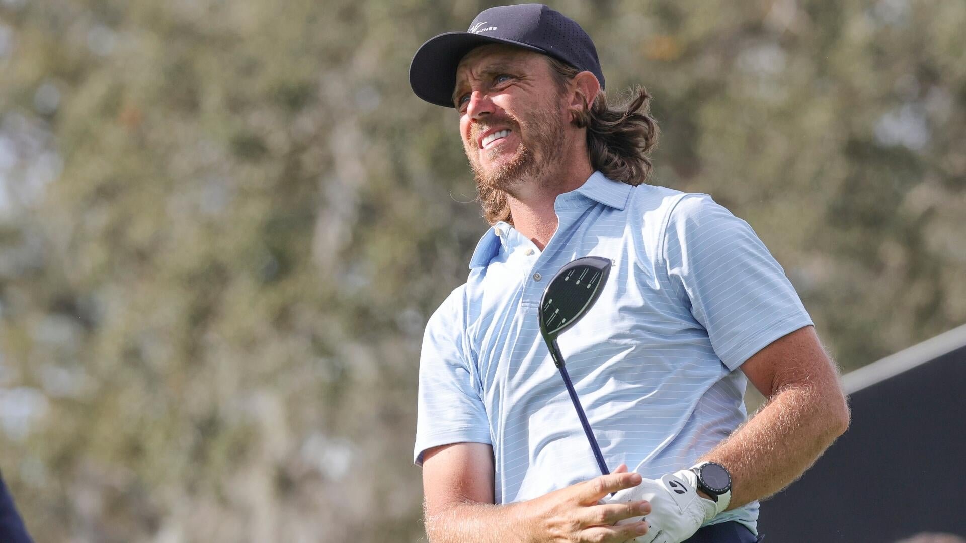 A bearded man in a navy cap and light blue polo shirt holds a golf club while looking upward, set against a blurred tree background.