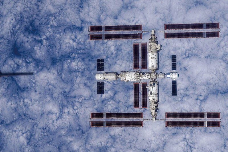 A space station with extended solar panels orbits above a dense layer of white clouds.