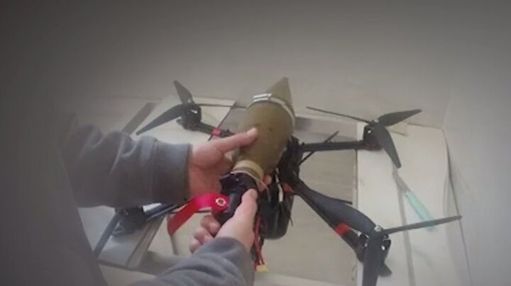 A person's hands attach an olive-green munition to a large black drone with multiple propellers, visible against a white backdrop.
