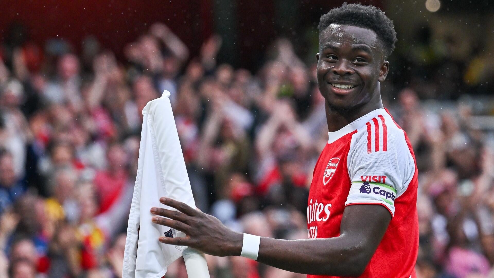 A smiling soccer player in a red and white Arsenal jersey with a captain's armband holds a white shirt while facing a cheering crowd.