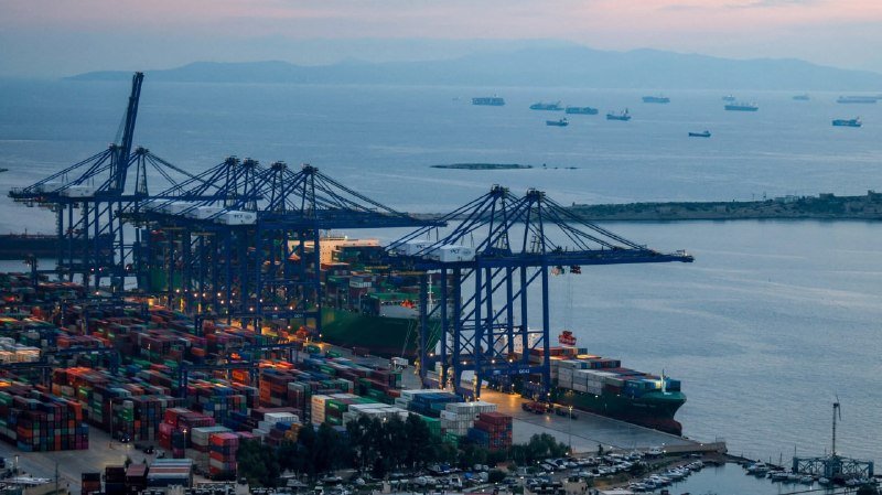 Large blue gantry cranes load containers onto a cargo ship at a port, with numerous shipping vessels anchored offshore under a hazy sky.