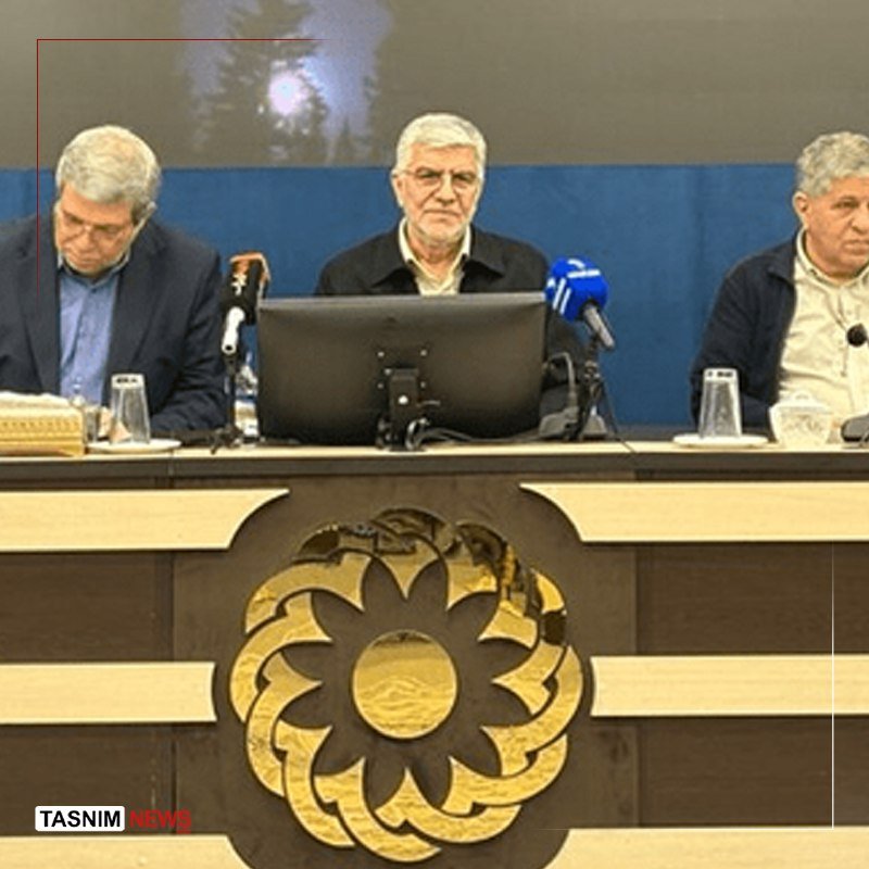 Three men sit at a panel table with microphones during a formal gathering, with a large golden emblem displayed on the desk's front panel.