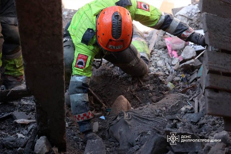 A rescuer in a high-visibility yellow uniform and orange helmet sifts through rubble and debris, reaching toward a body partially buried in the wreckage.