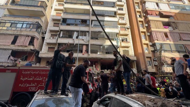 People gather around emergency responders and a red fire truck with Arabic writing outside a damaged apartment building.