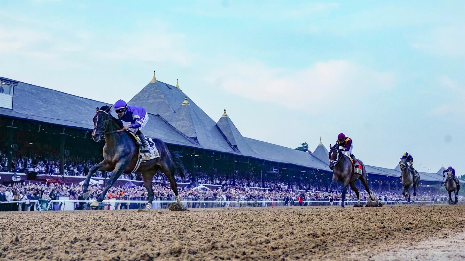 A jockey in purple silks rides a dark horse to the lead on a dirt track, with three other horses trailing behind, in front of packed grandstands and steeple-roofed buildings.