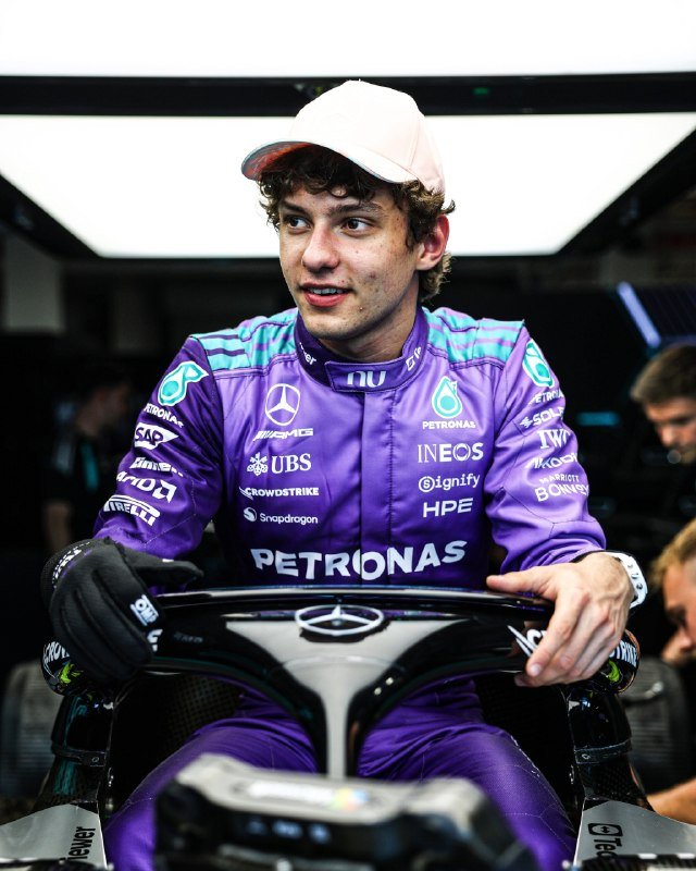 A young man wearing a purple Petronas-branded racing suit and pink cap sits in the cockpit of a black race car, looking off to the side.