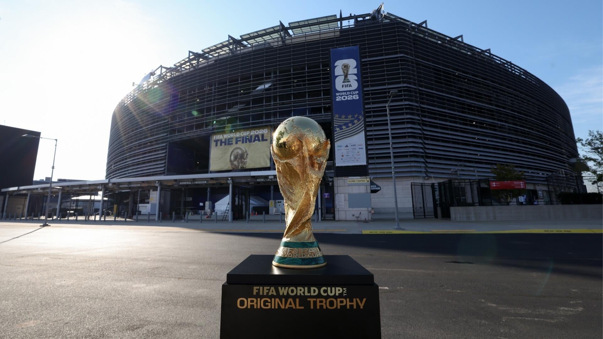 The FIFA World Cup Original Trophy sits on a black pedestal outside a stadium displaying banners reading "FIFA World Cup 2026 THE FINAL."