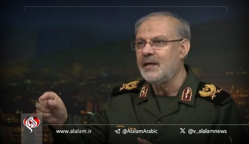 A bearded man in a military uniform with insignia gestures while speaking during an interview, with a nighttime cityscape backdrop and Al-Alam news network branding visible.