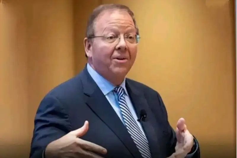 A man in a dark suit and striped tie gestures with both hands while speaking in front of a blurred tan background.