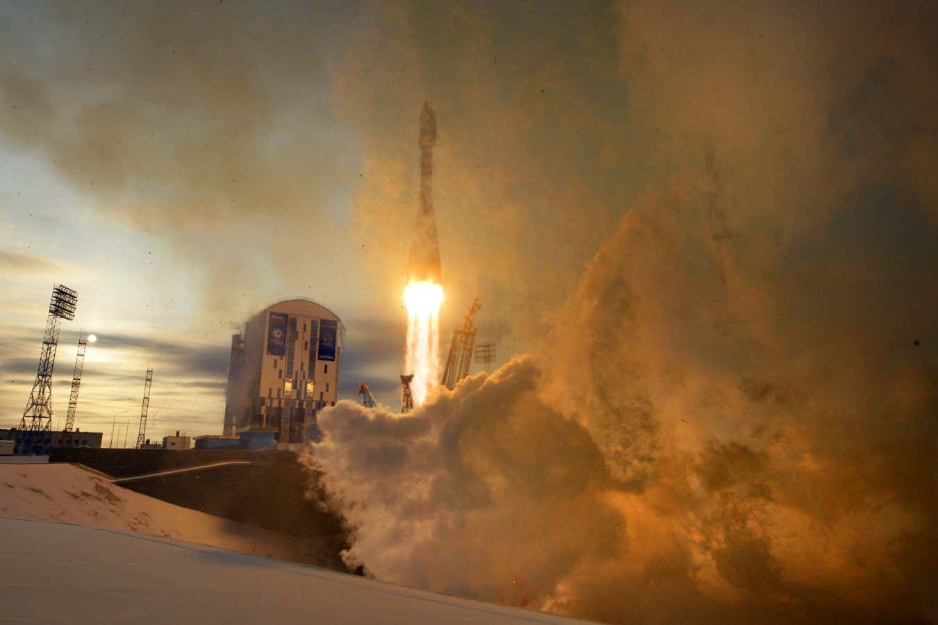 A rocket lifts off from a snowy launch pad, emitting bright flames and large plumes of smoke beside a service tower at dusk.
