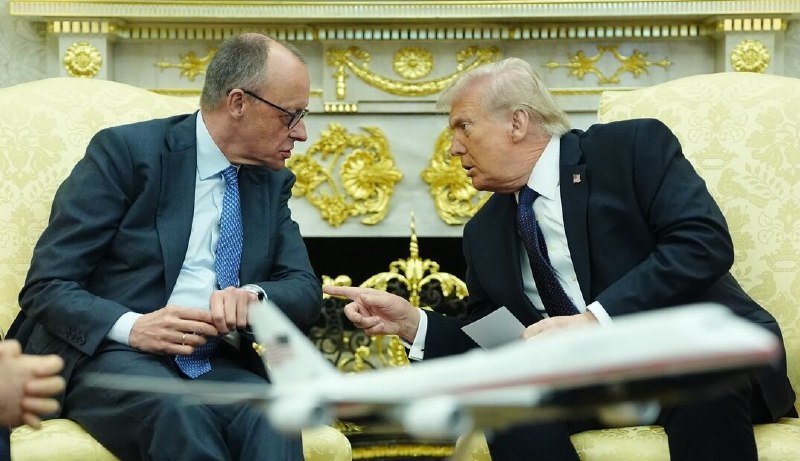 Two men in suits sit in ornate yellow chairs in a gilded room, leaning toward each other in conversation, with a model airplane blurred in the foreground.