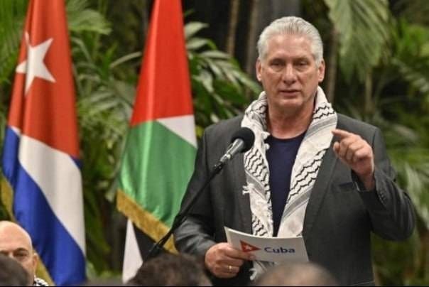 A man in a suit and keffiyeh scarf speaks at a podium, holding a paper labeled "Cuba," with Cuban and Palestinian flags visible behind him.