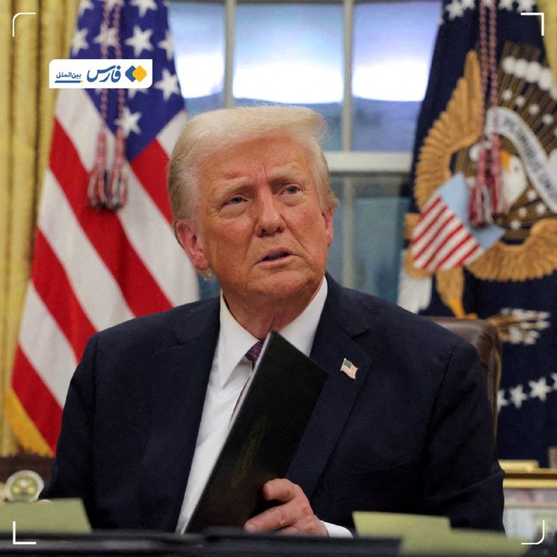 A man with blonde hair in a dark suit holds a black folder while seated in front of American flags.