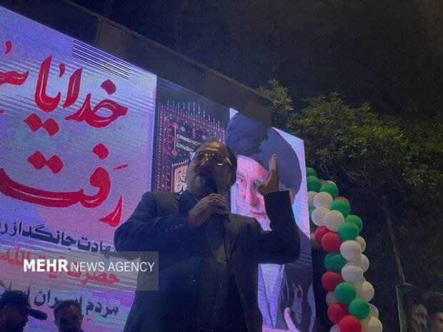 A bearded man in glasses speaks into a microphone at night, gesturing in front of a large banner with red Persian script and decorative balloons.