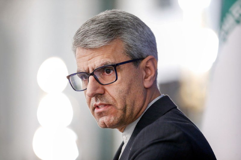 A gray-haired man in glasses and a dark suit looks off-camera against a blurred background with bright lights.