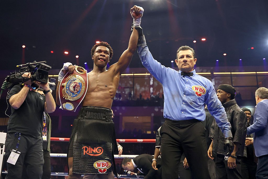 A shirtless boxer named "MASON" holds a championship belt while a referee in a WBO-branded shirt raises his arm in victory inside a boxing ring.