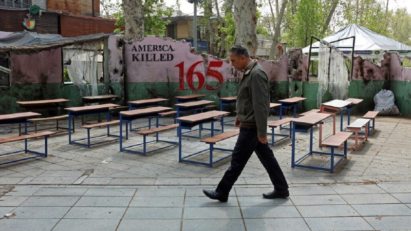 A man walks past empty school desks arranged outdoors in front of a pink wall reading "AMERICA KILLED 165."
