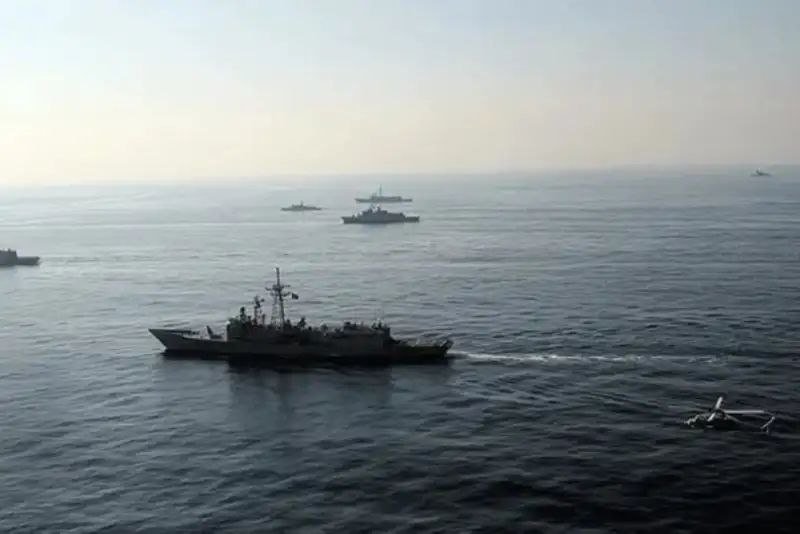 Multiple naval vessels sail across calm open water under a hazy sky, with a small boat floating in the foreground.