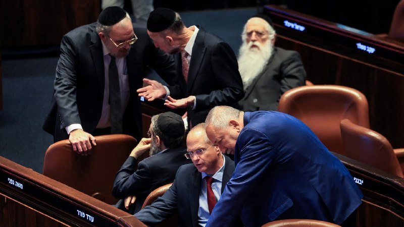 Several men in suits and kippot gather closely in conversation among rows of empty leather seats inside a wood-paneled chamber.