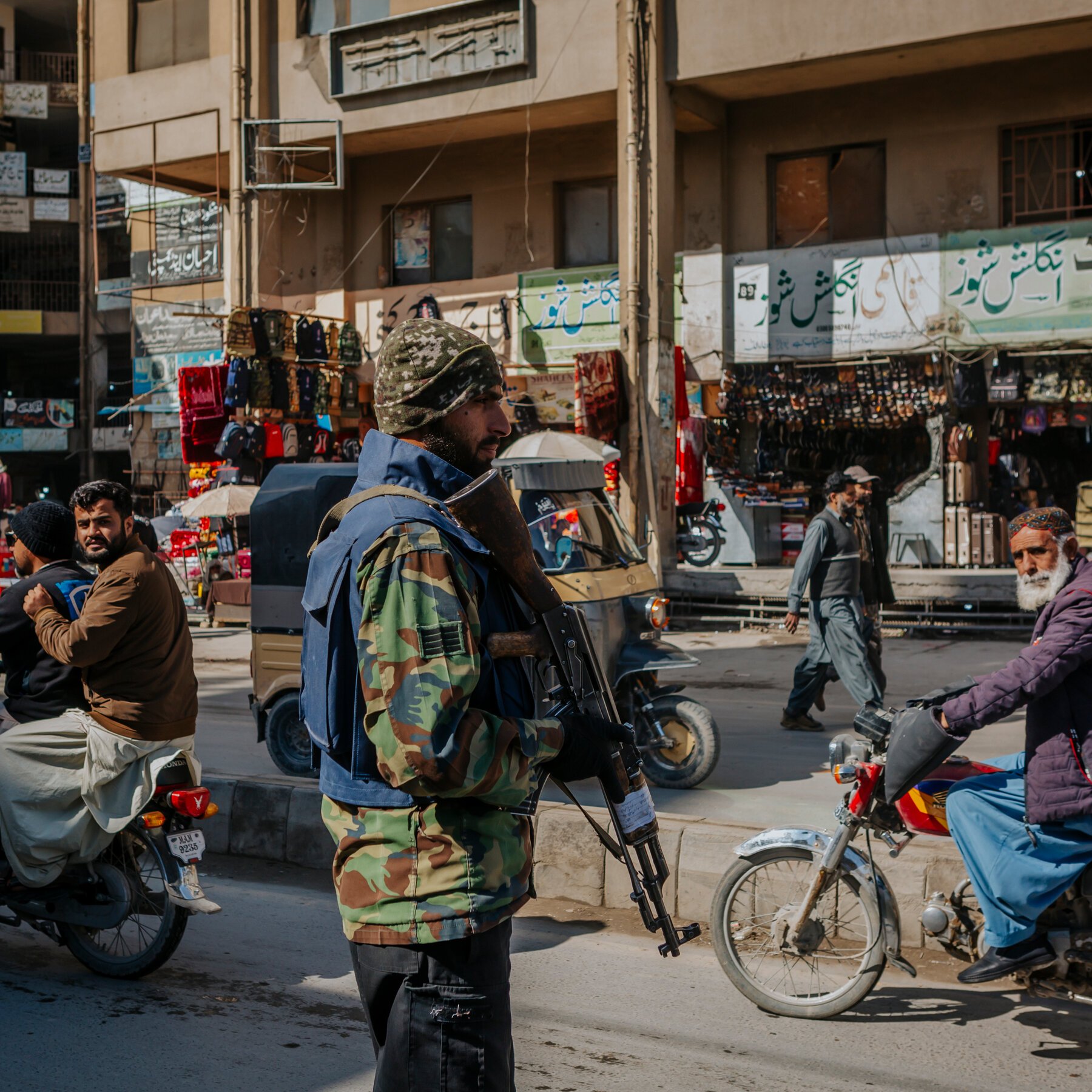 A bearded man in camouflage uniform and helmet stands holding a rifle on a busy street lined with shops bearing Urdu signage, while motorcyclists and pedestrians pass by.