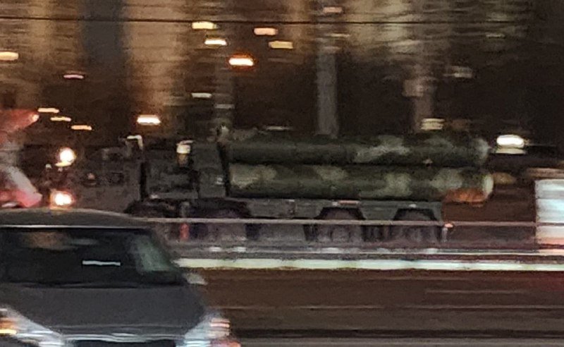 A camouflage-painted missile launcher mounted on a truck is transported along a city street at night, with a car visible in the foreground.