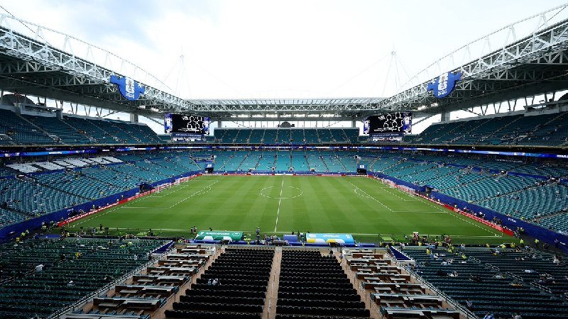 A large, nearly empty stadium with a green soccer pitch, surrounded by blue seating tiers and large digital screens displaying event graphics.