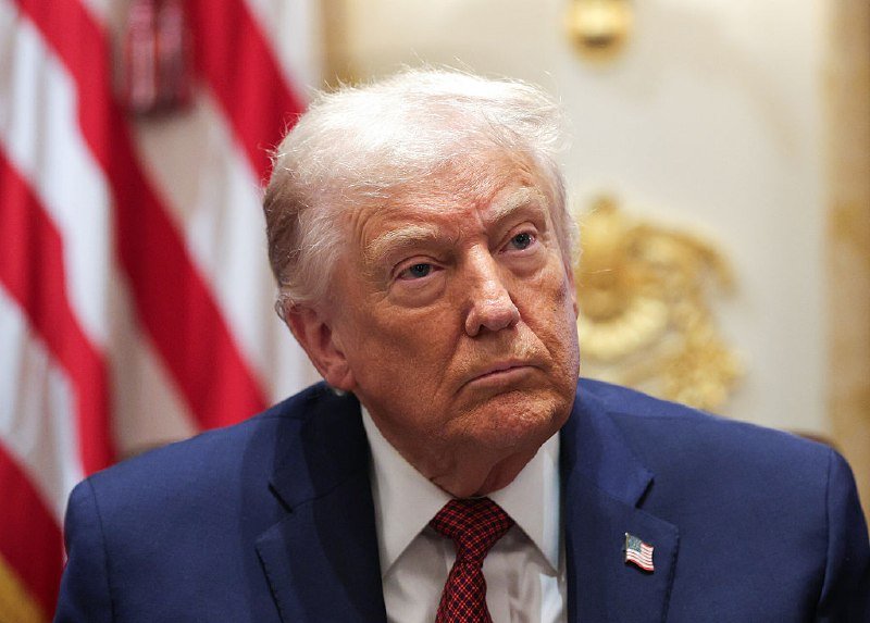 An older man with white hair, wearing a dark blue suit, white shirt, and red patterned tie with an American flag pin, sits in front of a U.S. flag and ornate gold decor.