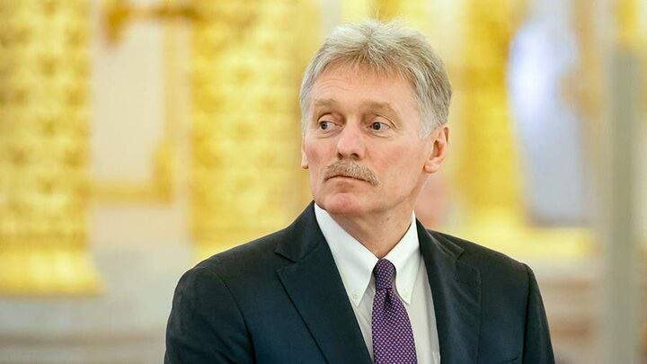 A gray-haired man with a mustache, wearing a dark suit and purple tie, looks to the side against a blurred gold-background setting.