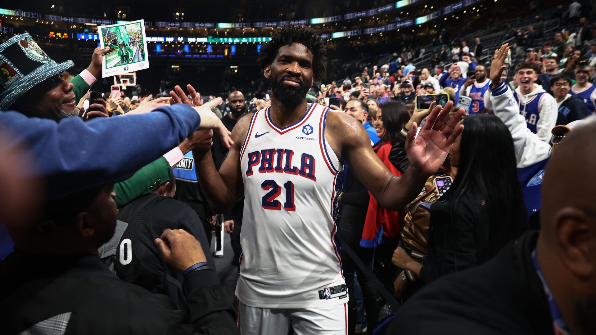 A bearded basketball player wearing a white "PHILA 21" jersey smiles while waving to fans reaching out and recording him with phones inside an arena.