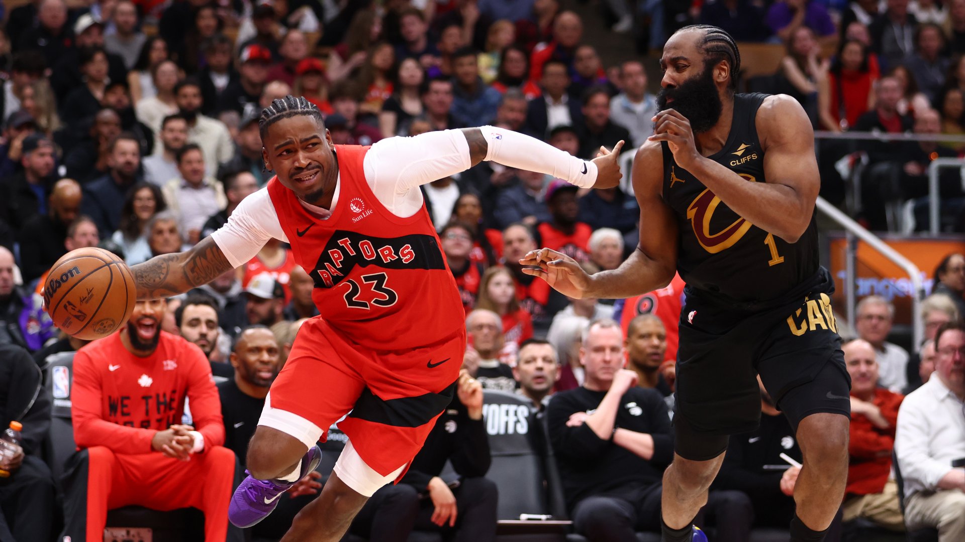 A basketball player in a red Raptors jersey numbered 23 dribbles a ball while being defended by a player in a black Cavaliers jersey numbered 1, before a crowded arena audience.