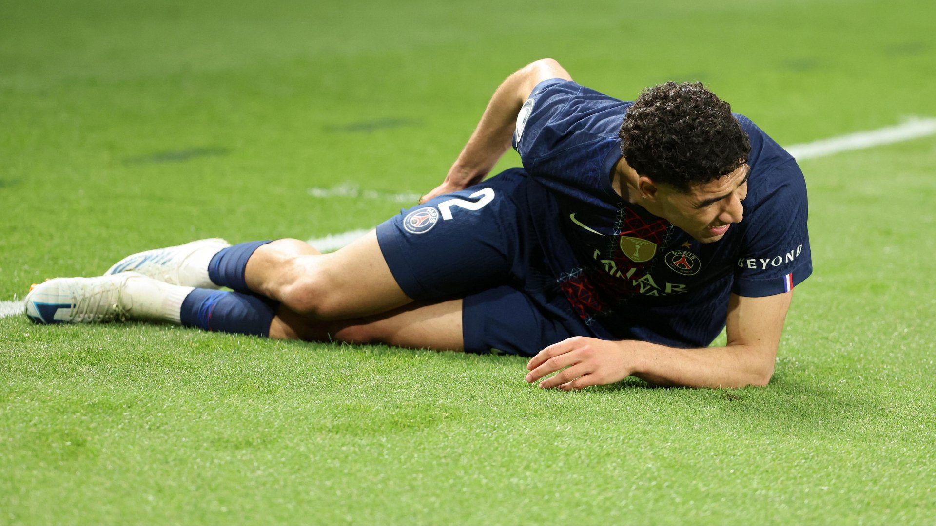 A soccer player in a dark blue PSG jersey lies on the grass field, appearing to grimace while holding his side.