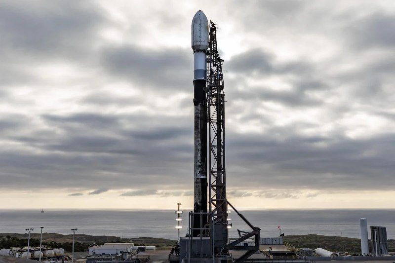 A two-stage rocket with "SpaceX" and "Falcon 9" markings stands vertically on a coastal launch pad beside a support tower under a cloudy sky.