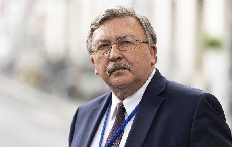 An older man with glasses, a mustache, a dark suit, and a blue lanyard stands outdoors looking toward the camera.