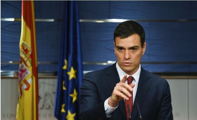 A dark-haired man in a navy suit and red tie gestures with his hand in front of the Spanish and European Union flags.