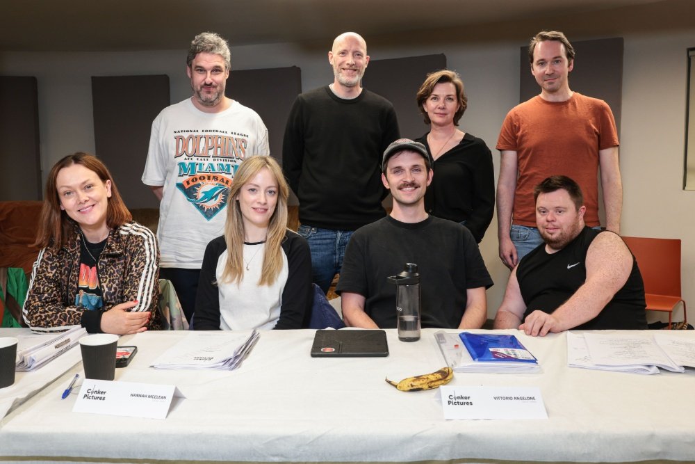 Eight people pose behind a table with scripts, a water bottle, a banana, and name cards reading "Hannah McLean" and "Vittorio Angelone."