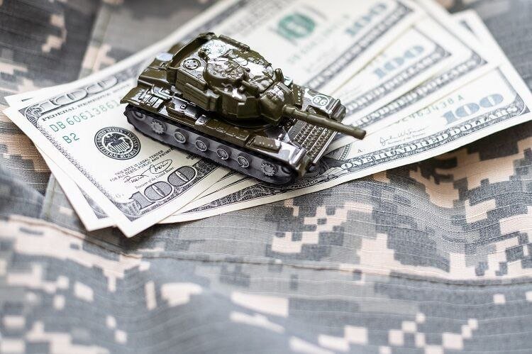 A small toy tank rests on a fan of U.S. one-hundred-dollar bills spread across a camouflage-patterned fabric surface.