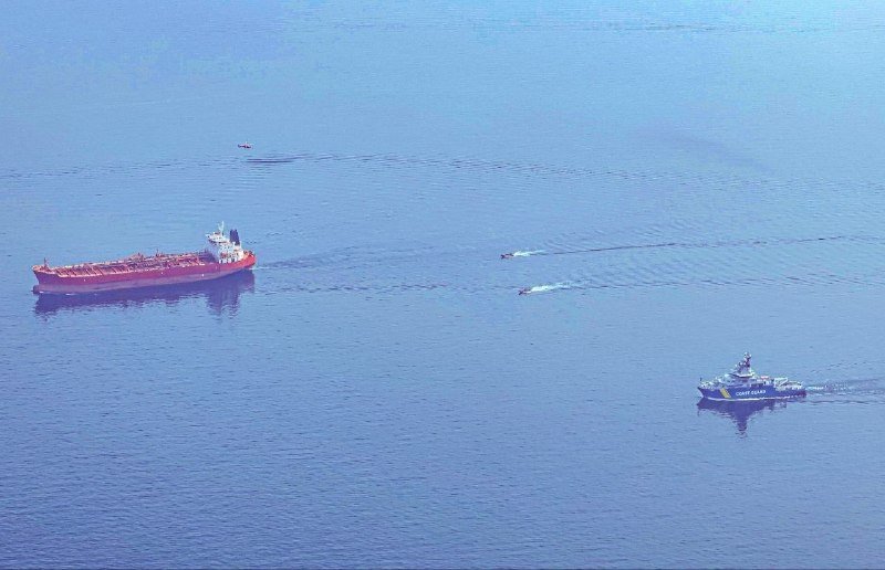 An aerial view shows a red cargo ship and a blue-and-white coast guard vessel navigating through blue waters, with smaller boats visible in the distance.