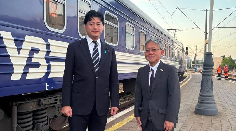 Two men in business suits stand smiling on a railway platform in front of a blue and white passenger train.