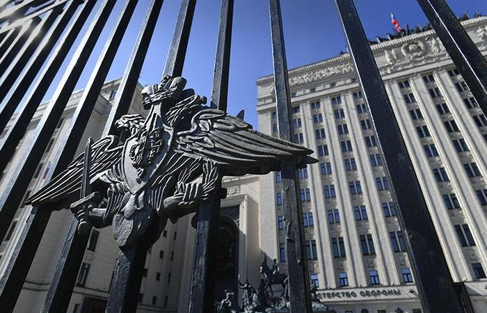 A metal emblem featuring a double-headed eagle is mounted on black iron gate bars, with a tall, light-colored government building displaying a red flag visible in the background under a clear blue sky.