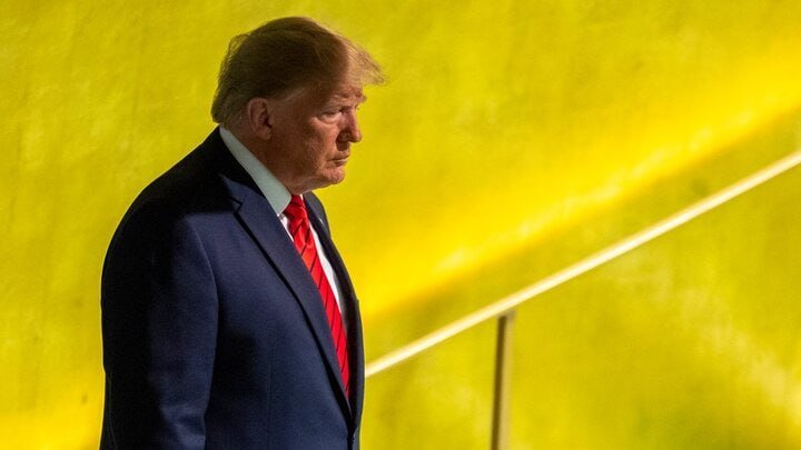 A man with blond hair in a dark suit and red tie walks past a yellow wall.