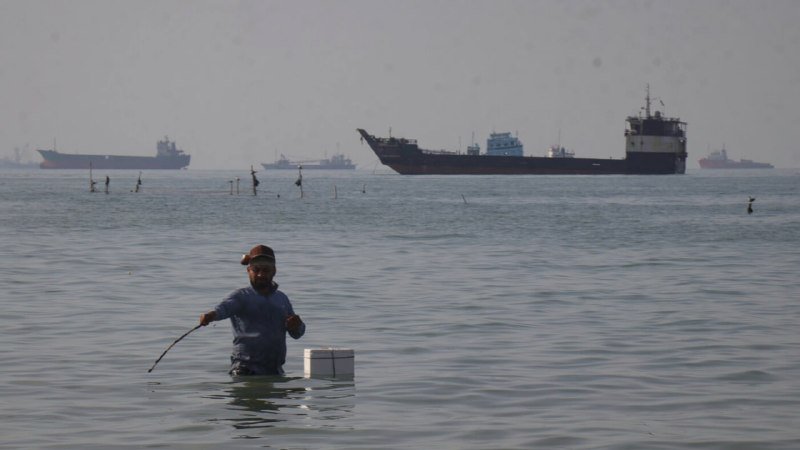 A man stands waist-deep in water holding a long tool, with a small cooler floating nearby, as large cargo ships sit anchored in the hazy background.