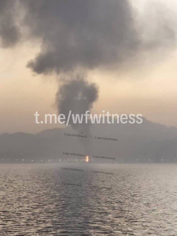 A large plume of dark smoke rises above a body of water at dusk, with hills and faint lights visible in the distance.