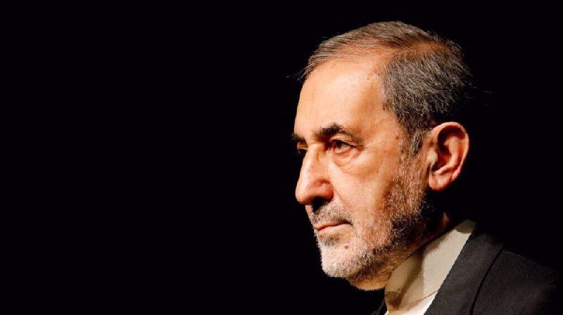 A gray-bearded man in a dark suit is shown in profile against a black background, gazing toward the left.