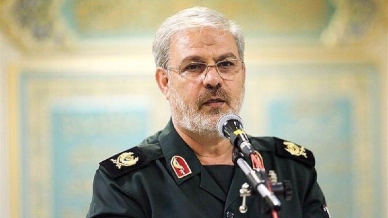 A gray-bearded man in a dark green military uniform with insignia speaks into a microphone during an indoor event.