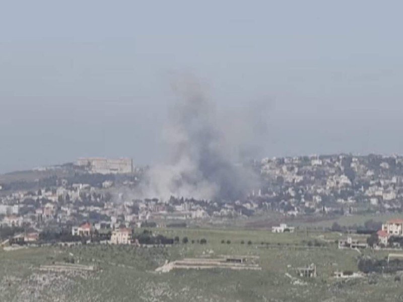 A large plume of gray smoke rises from a hillside town surrounded by green fields under a hazy sky.