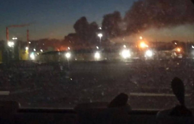 A blurry nighttime view shows a large crowd, distant bright lights, and a plume of smoke rising against a dark sky.