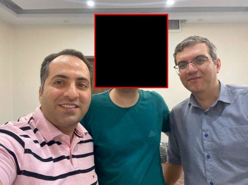 Three men pose for a close-up photo indoors, with the middle person's face obscured by a black square outlined in red.