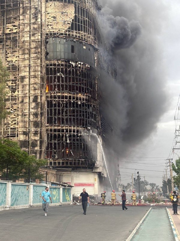 Firefighters spray water on a charred, multi-story building engulfed in thick black smoke, while bystanders and emergency responders gather on the street below.