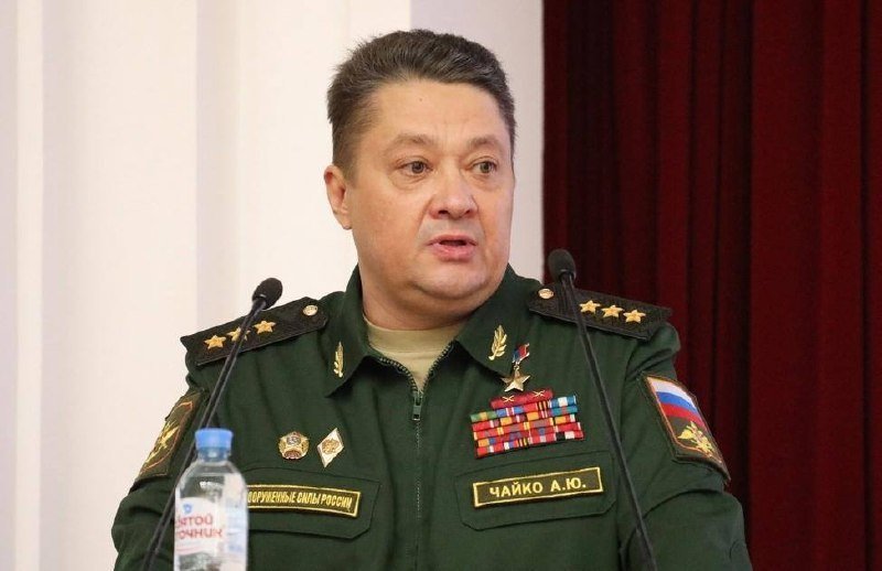 A Russian military officer in a green uniform with four stars and medal ribbons speaks at a podium with microphones, a water bottle, and red curtains in the background.