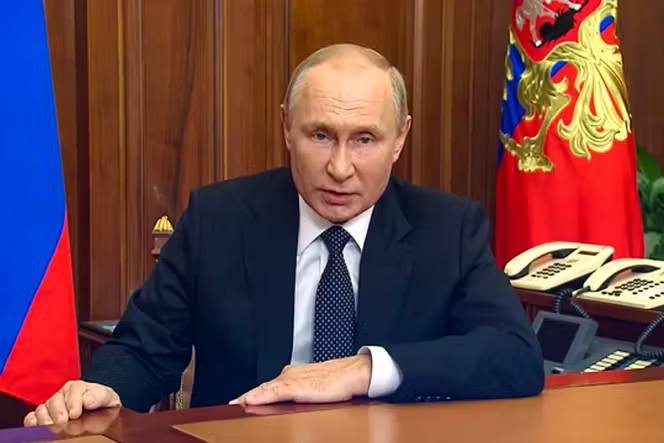 A man in a dark suit and dotted tie sits at a wooden desk speaking, with a tricolor and a red flag bearing a gold emblem behind him.